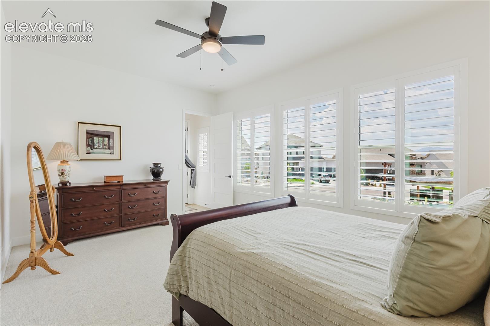 White walls, ceiling fan, carpet flooring, multiple windows with plantation shutters, and a white interior door
