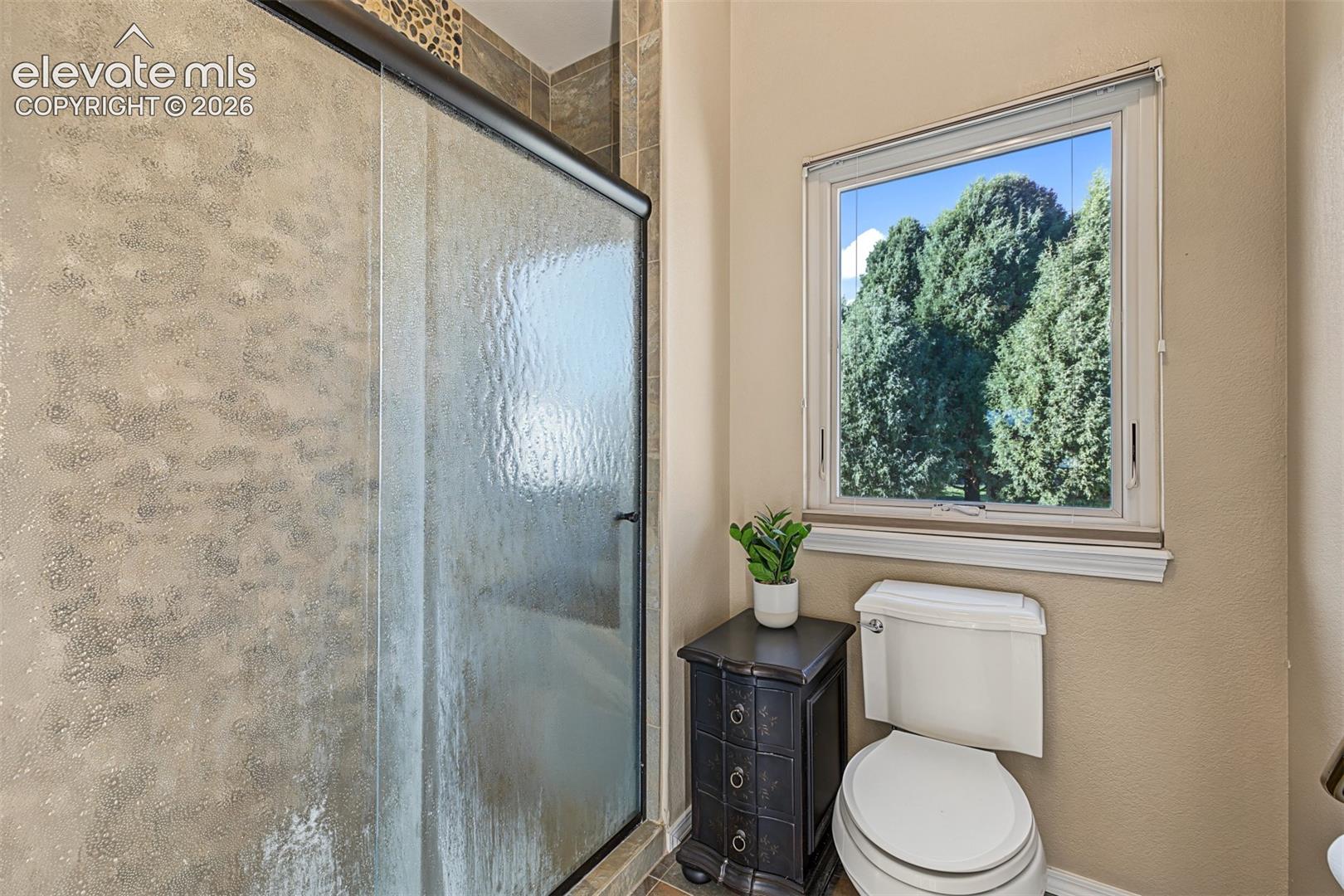 Additional view of the primary bathroom featuring a walk-in shower with textured glass enclosure, tile surround, and a large window providing natural light and views of mature trees.