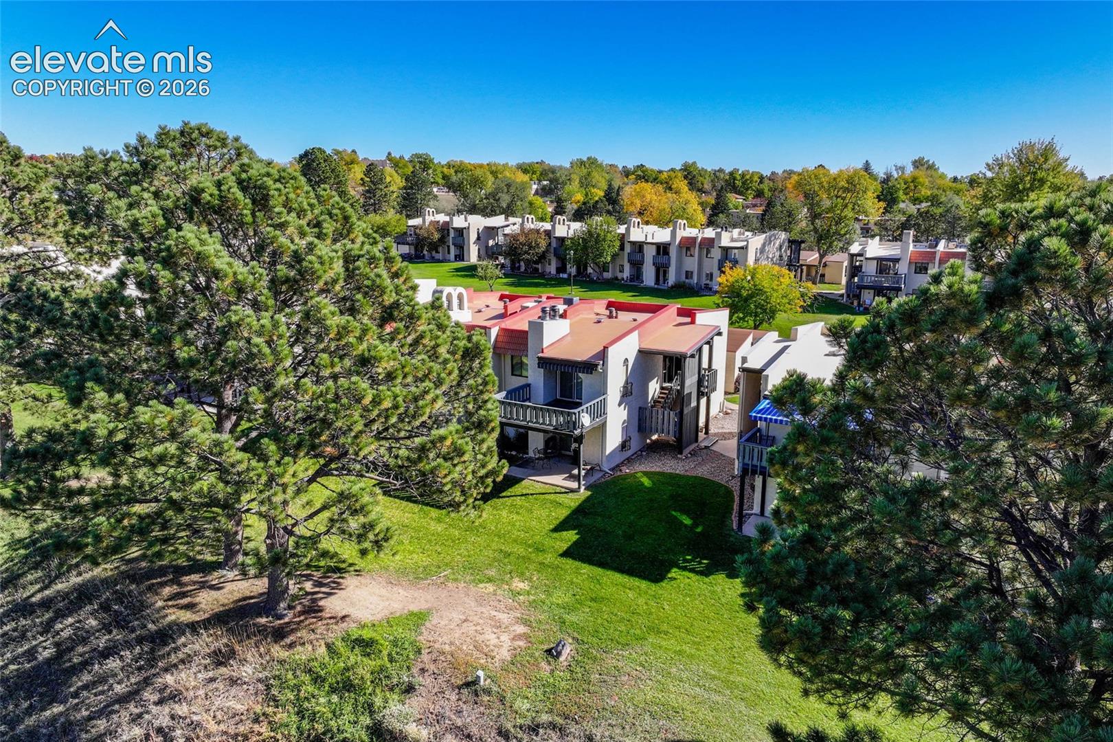 Aerial perspective featuring multi-story residential buildings with metallic roofing, stucco exteriors, attached balconies, and expansive surrounding lawns with mature pine trees