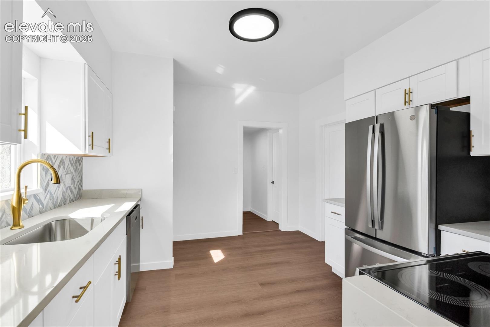 Kitchen featuring white shaker-style cabinetry with gold-tone hardware, wood-finish flooring, stainless steel appliances, and a herringbone-patterned tile backsplash