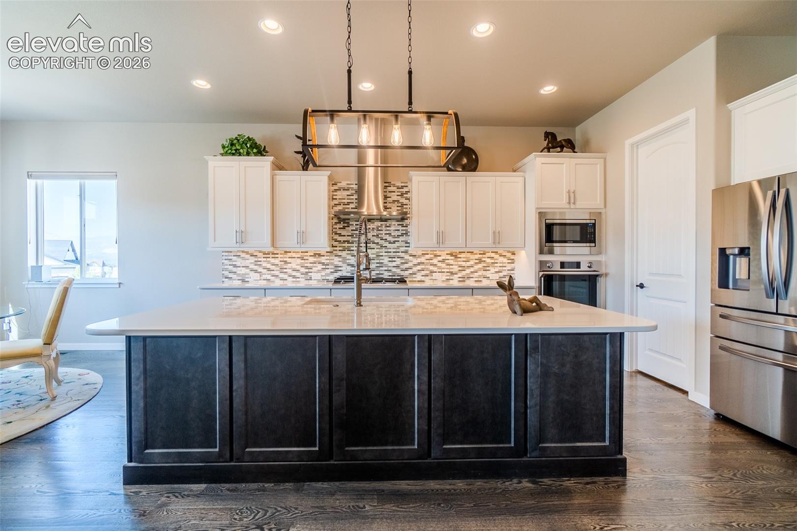 Kitchen featuring an expansive center island with a light-toned countertop, a stylish linear pendant light fixture, stainless steel appliances, a mosaic tile backsplash, and wood-finish flooring