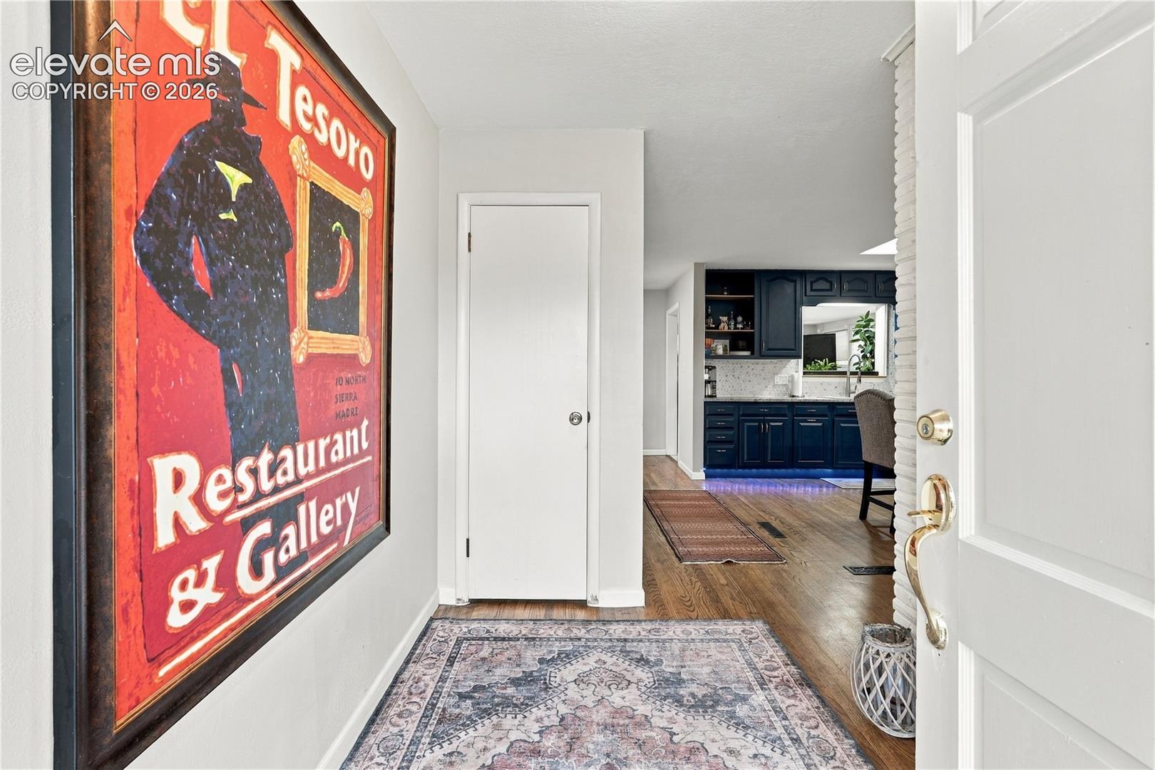 Entryway featuring wood-finish flooring and a white paneled door, revealing a kitchen with dark cabinetry, tile backsplash, and a pass-through opening