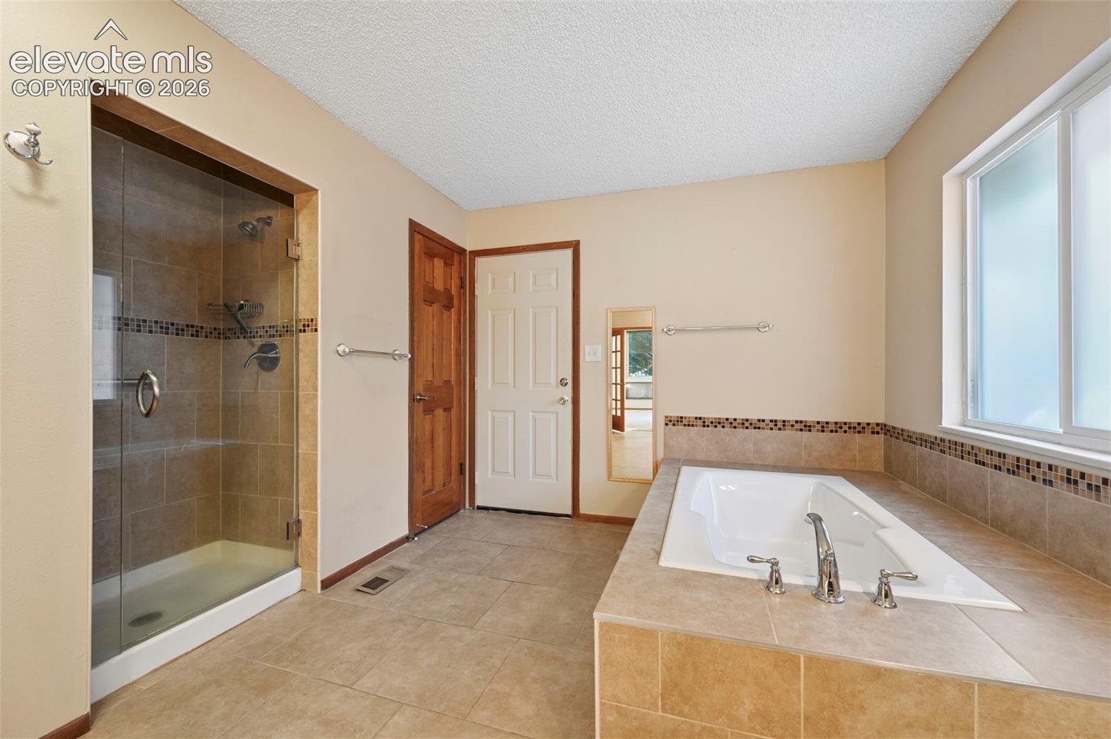 Bathroom featuring a tiled walk-in shower with a glass door, a built-in soaking tub with tile surround, decorative mosaic accent tiling, ceramic floor tile, and wooden door finishes