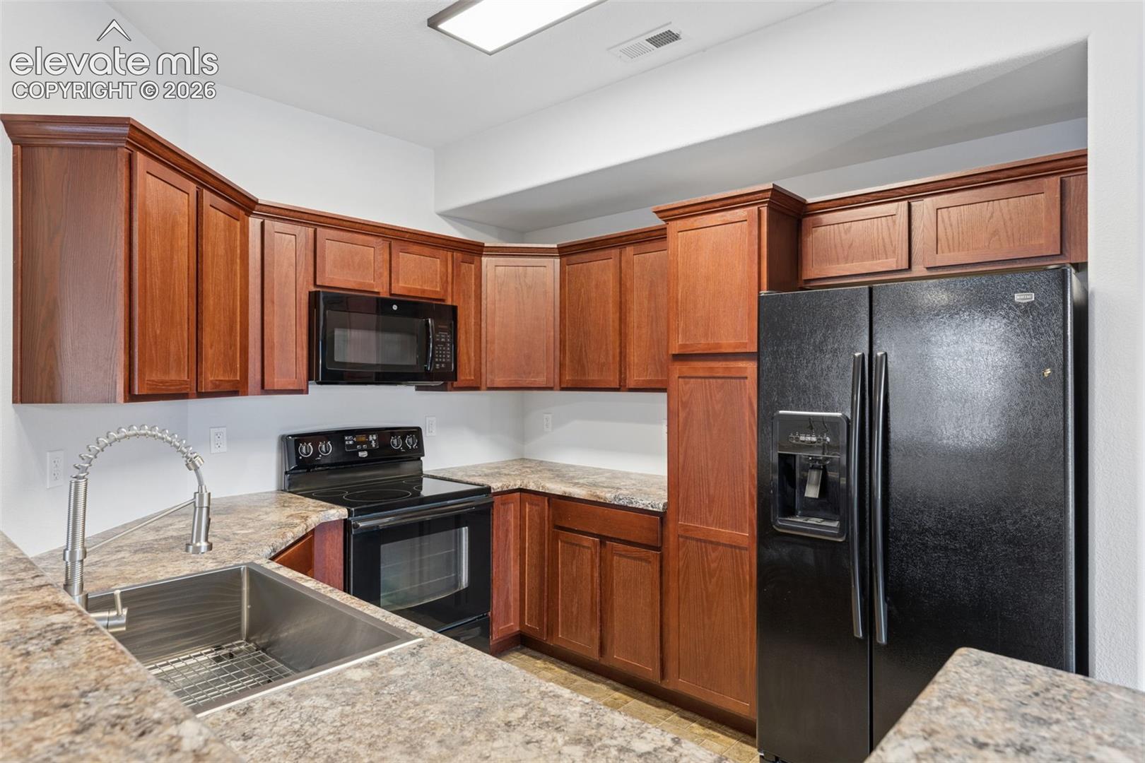 Kitchen featuring wood cabinetry, stone-finish countertops, stainless steel single-basin sink with arched pull-down faucet, and black appliance suite including a built-in microwave and refrigerator