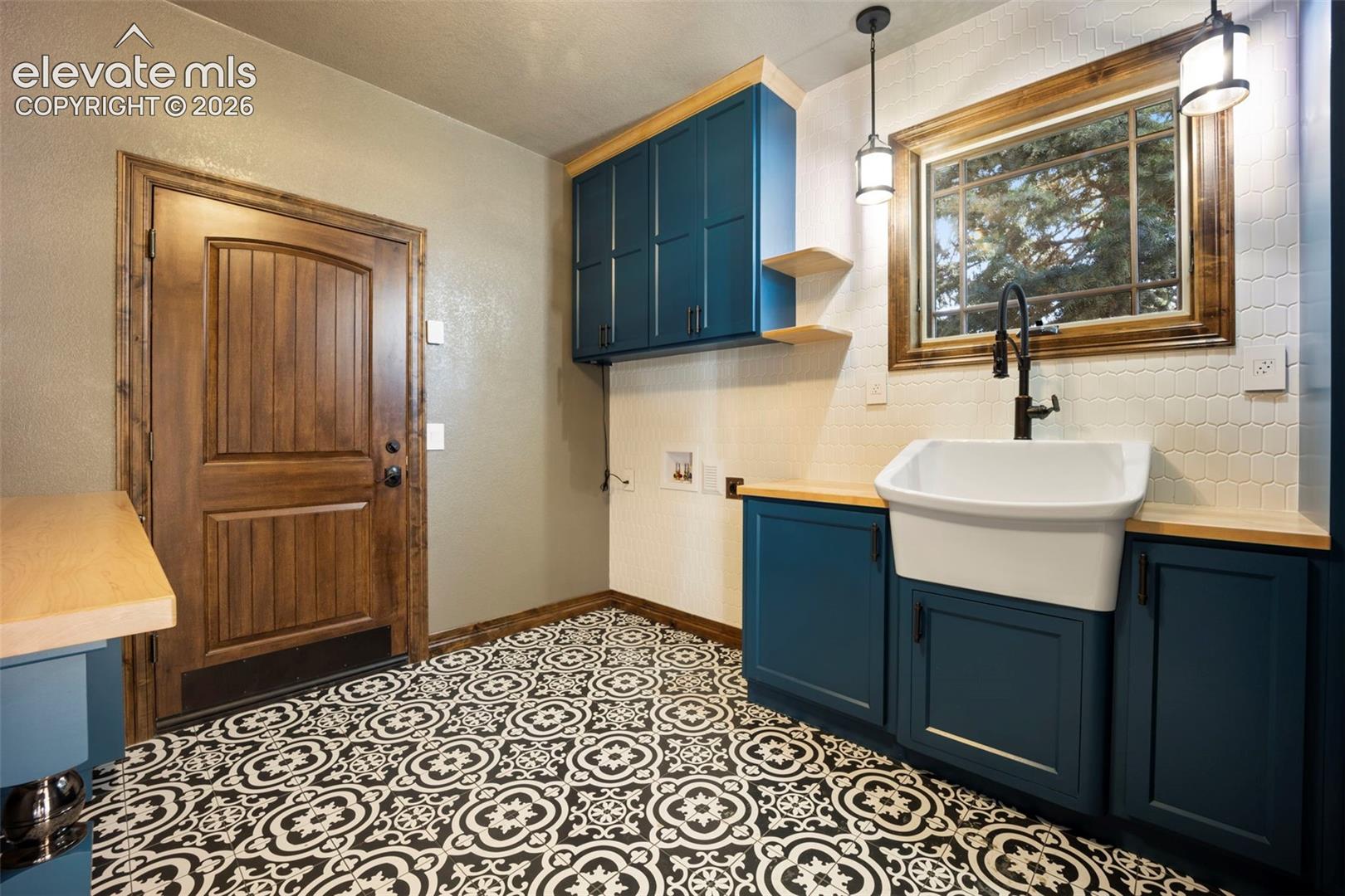 Utility space featuring patterned floor tiles, a deep apron-front sink with a black gooseneck faucet, teal cabinetry with wood-finish countertops, and a wood-framed window with pendant lighting