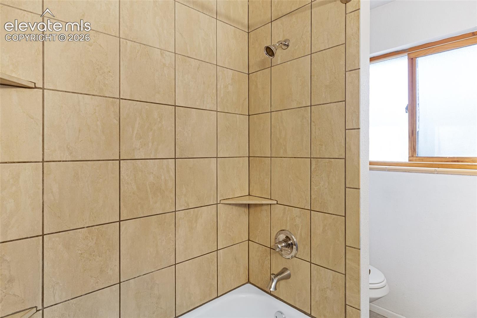 Tiled shower enclosure featuring neutral stone-finish ceramic tiles, chrome showerhead and trim, built-in corner shelf, and bathtub with integrated surround