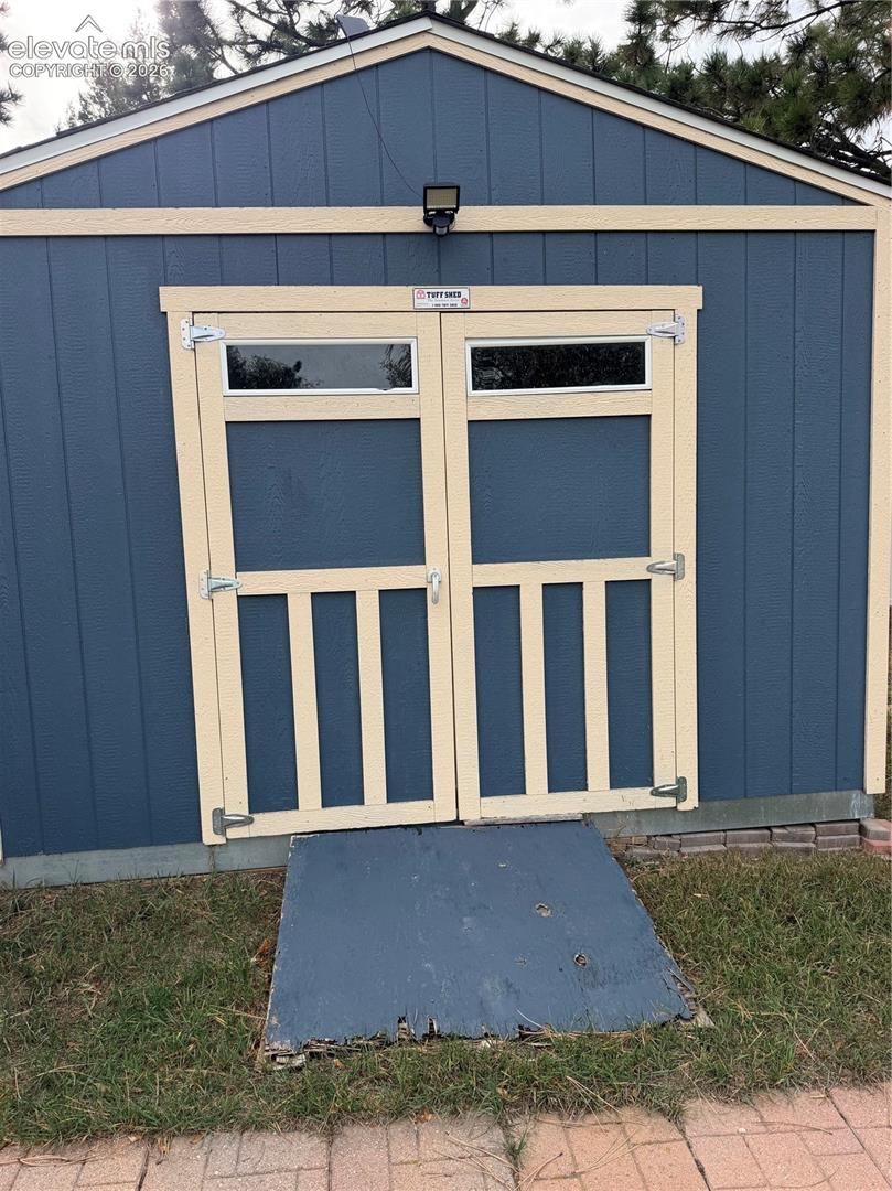Storage shed featuring vertical-panel siding, double doors with upper transom windows, a dedicated exterior lighting fixture, and an integrated ramp entry