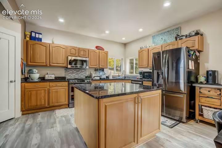 Kitchen featuring a central island with a dark countertop, stainless steel appliances, light wood cabinetry, a tiled backsplash, and wood-finish flooring