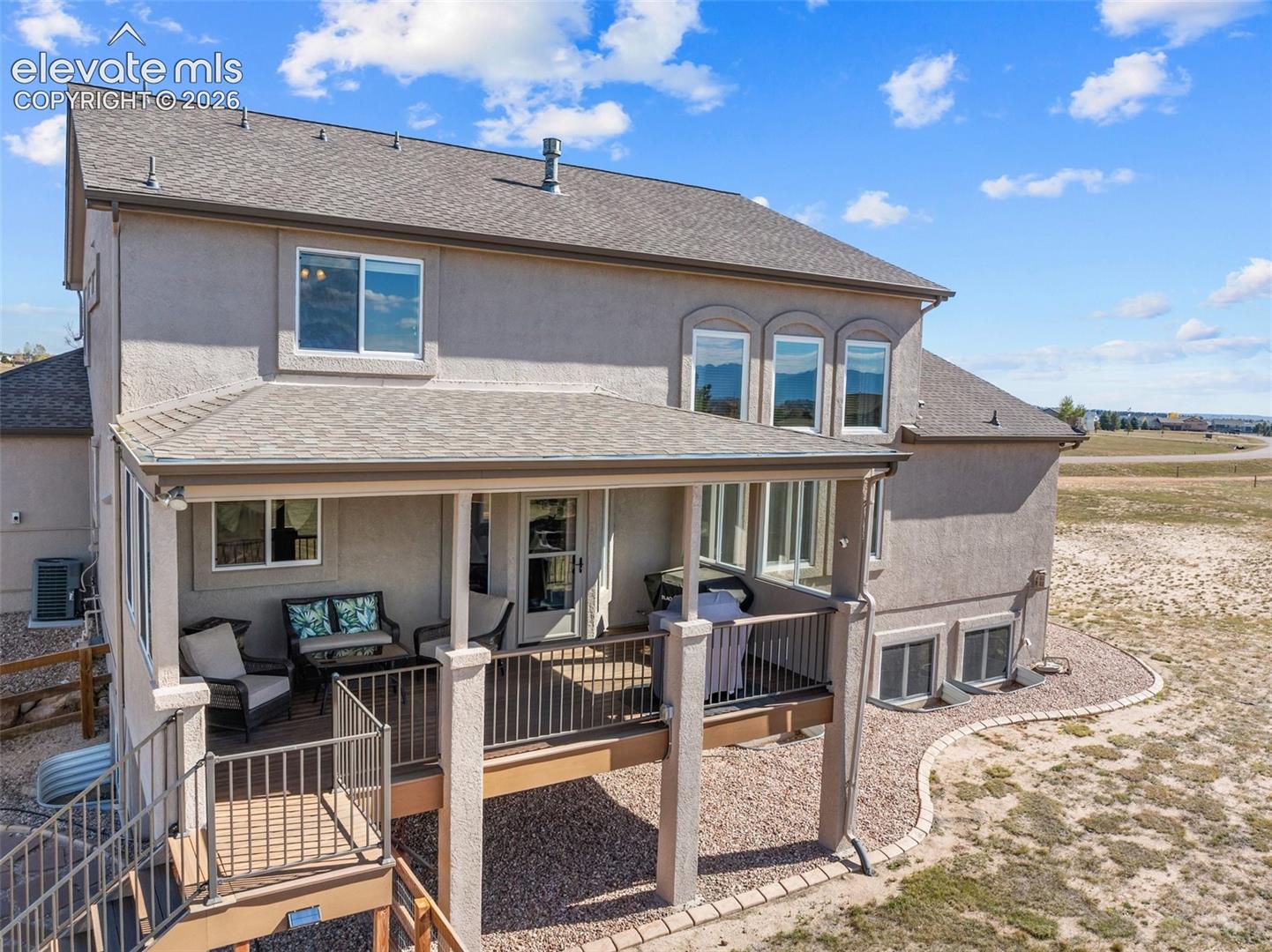 Stucco exterior with a covered rear deck featuring metal railings, support columns, and stairs leading to a graveled backyard