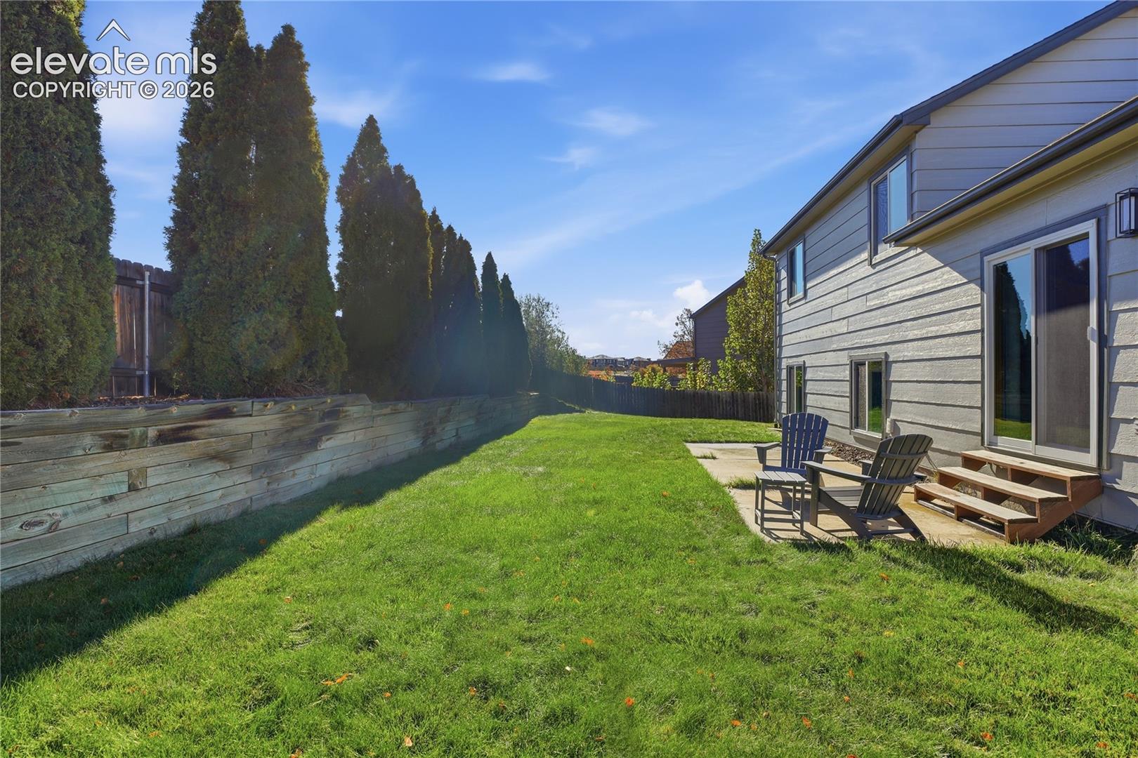 REAR EXTERIOR FEATURING HORIZONTAL LAP SIDING, A CONCRETE PATIO, WOODEN LANDSCAPE TIMBER RETAINING WALL, AND MATURE EVERGREEN BORDER TREES