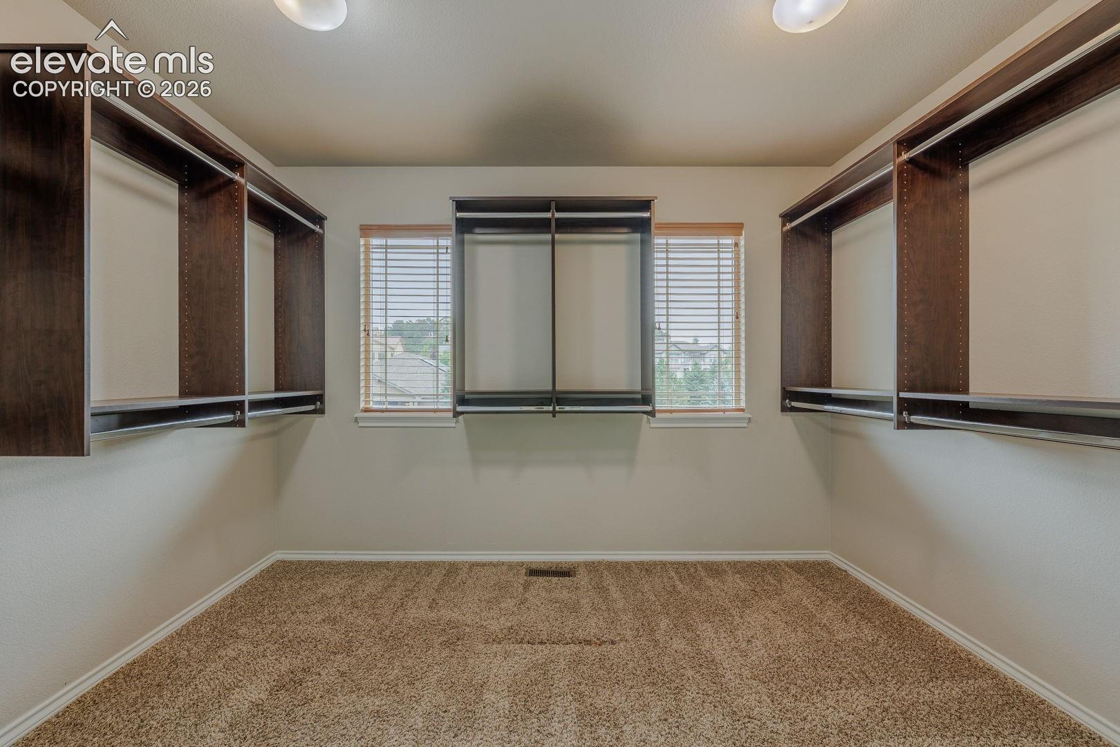Walk-in main bedroom closet featuring built-in wood-finish shelving, multiple hanging rods, and twin windows with blinds