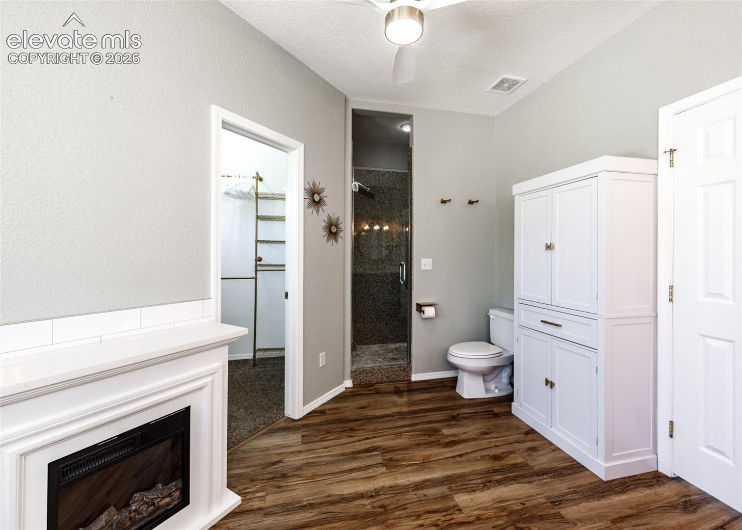 Electric fireplace with white mantle, wood-finish flooring, built-in white cabinetry, and recessed shower stall with tiled surround