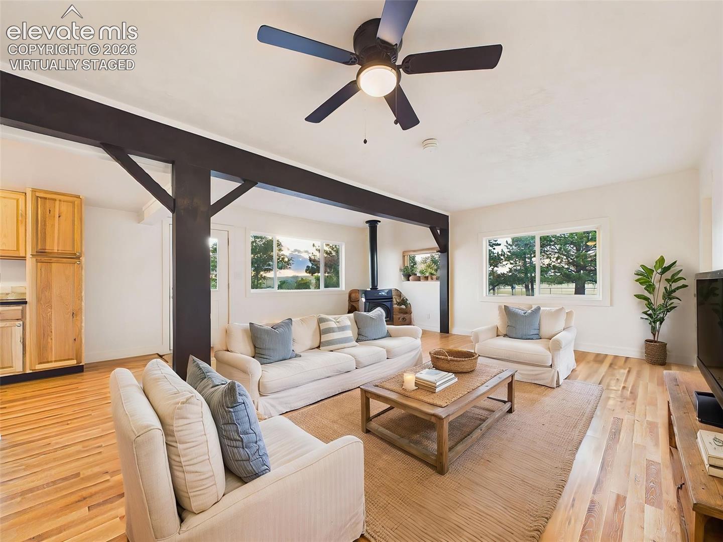 (Virtually Staged) Great Room – Spacious great room featuring exposed beams, abundant natural light, and a freestanding wood-burning fireplace that creates a warm focal point.