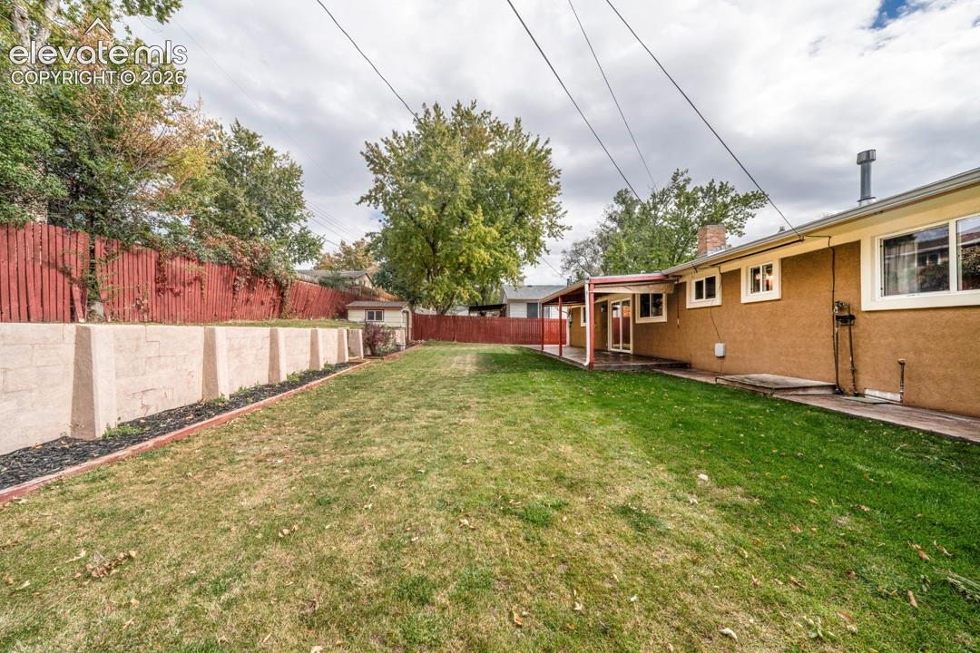 Fenced backyard featuring a manicured lawn, concrete retaining wall, covered patio, and storage shed