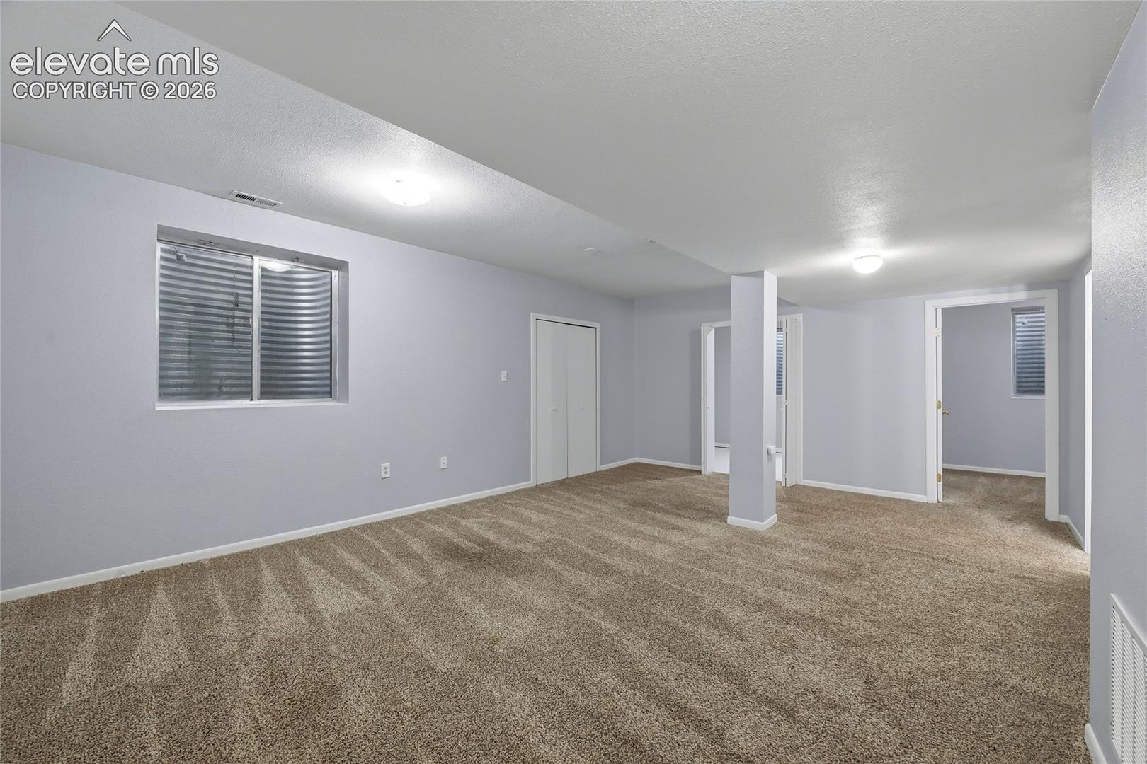 Interior room featuring neutral-toned carpeting, flush-mount ceiling lighting, inset egress window with metal window well, and bi-fold closet doors