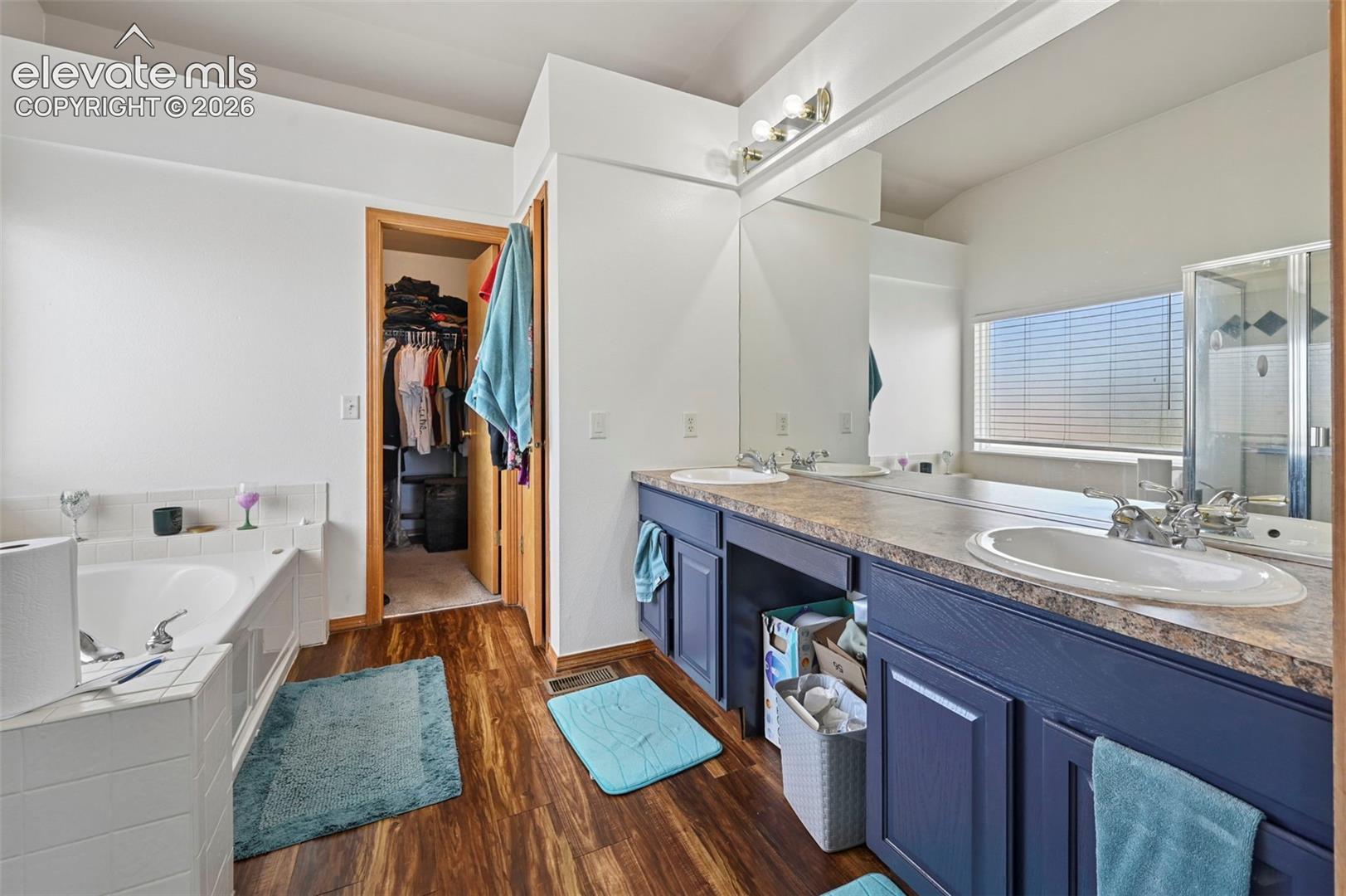Bathroom featuring wood-finish flooring, a built-in tub with white tile surround, a double vanity with dark blue cabinetry, and a walk-in shower with clear glass enclosure