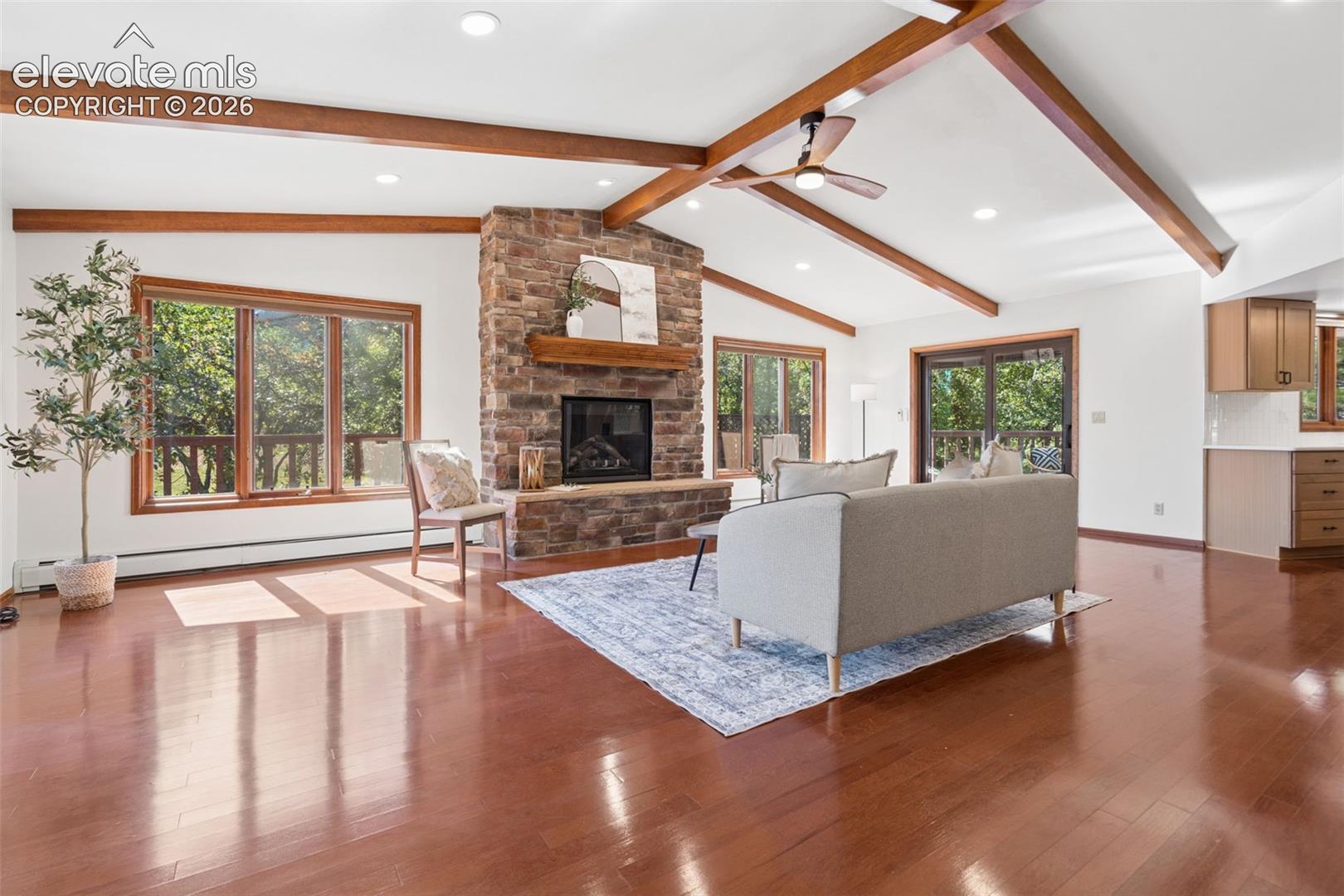 Vaulted ceiling with exposed wood beams, stone fireplace, wood-finish flooring, recessed lighting, and a ceiling fan