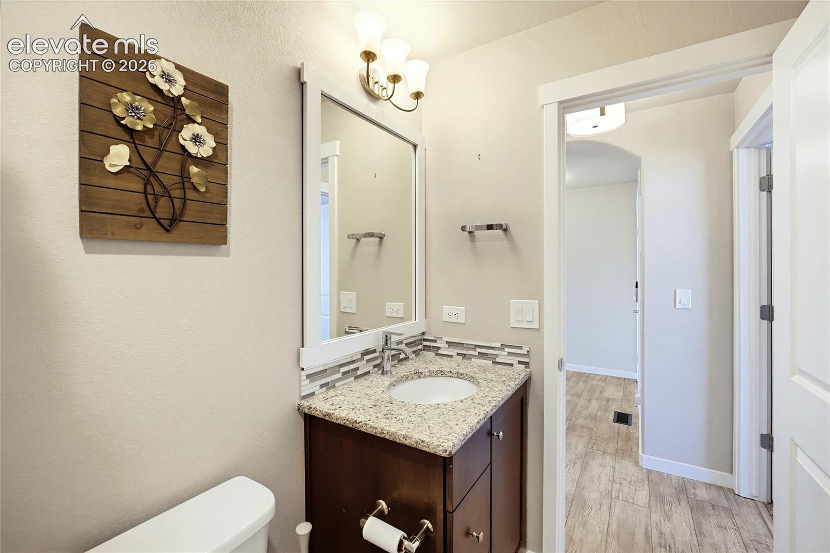Vanity featuring stone-style countertop, undermount sink, multi-tone mosaic tile backsplash, dark wood-finish cabinetry, and three-light wall-mounted fixture
