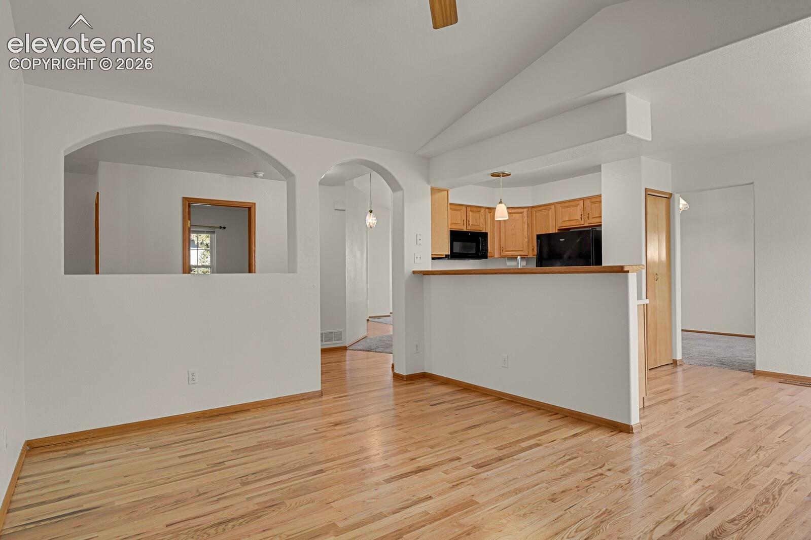 Vaulted ceiling, wood-finish flooring, kitchen with wood cabinetry and black appliances, arched wall openings, and pendant lighting