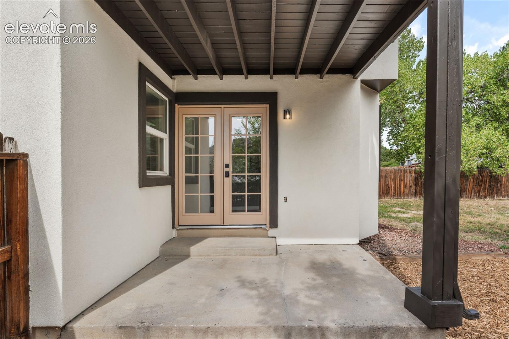 Covered patio featuring a concrete slab, wood-beamed ceiling, structural post, dual-paned glass entry doors, and wall-mounted exterior lighting