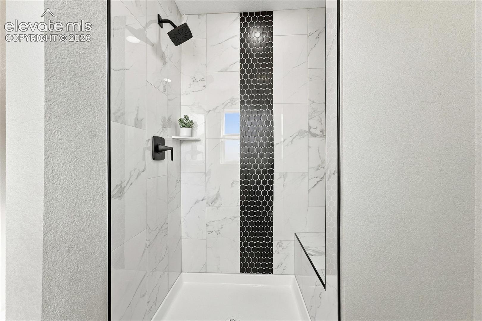 Walk-in shower featuring large-format white tile with gray veining, a central vertical black hexagonal tile accent, a built-in bench, matte black plumbing fixtures, and a glass surround