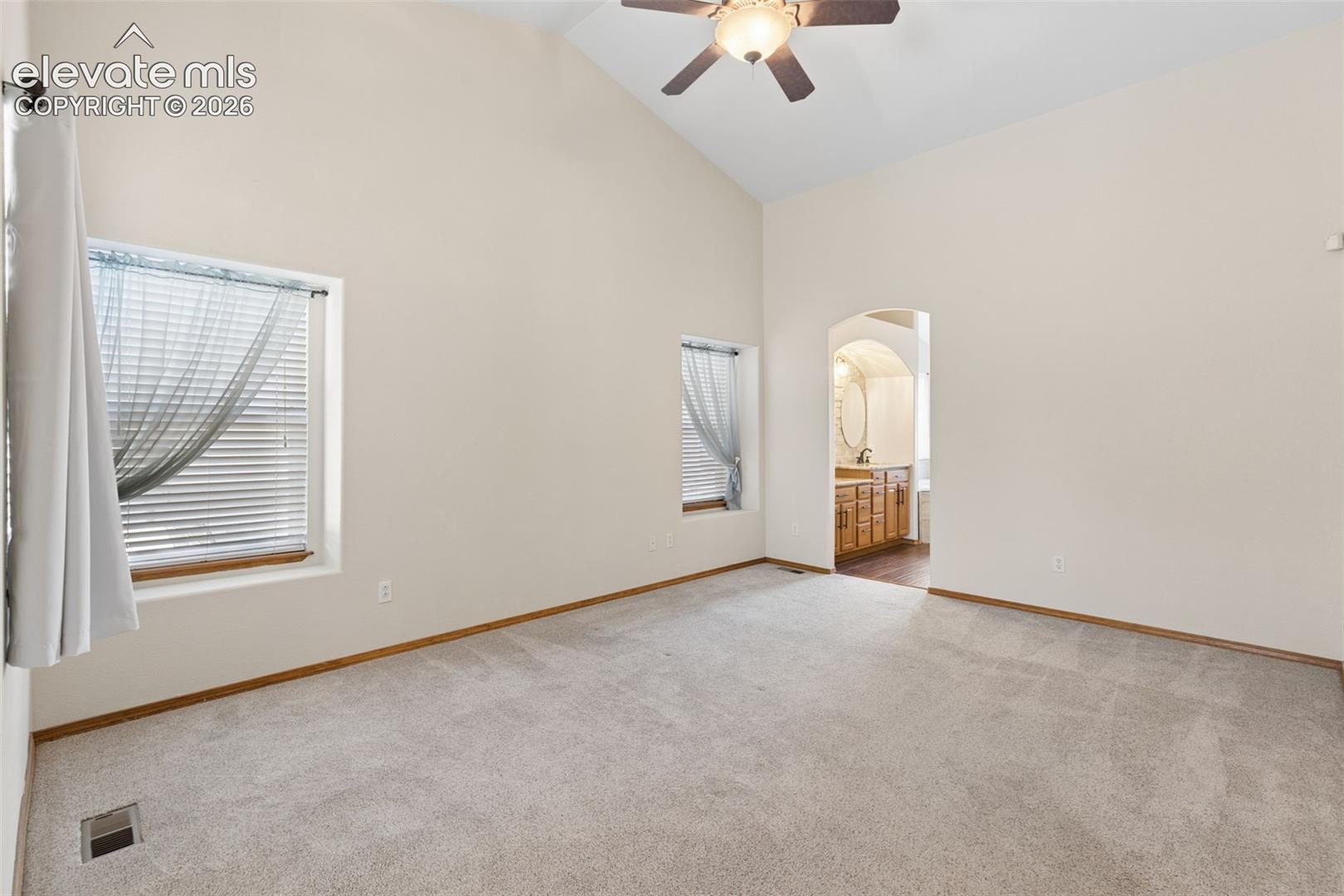 Vaulted ceilings featuring a central ceiling fan, neutral wall-to-wall carpeting, two recessed windows, and an arched doorway leading to a vanity area with wooden cabinetry