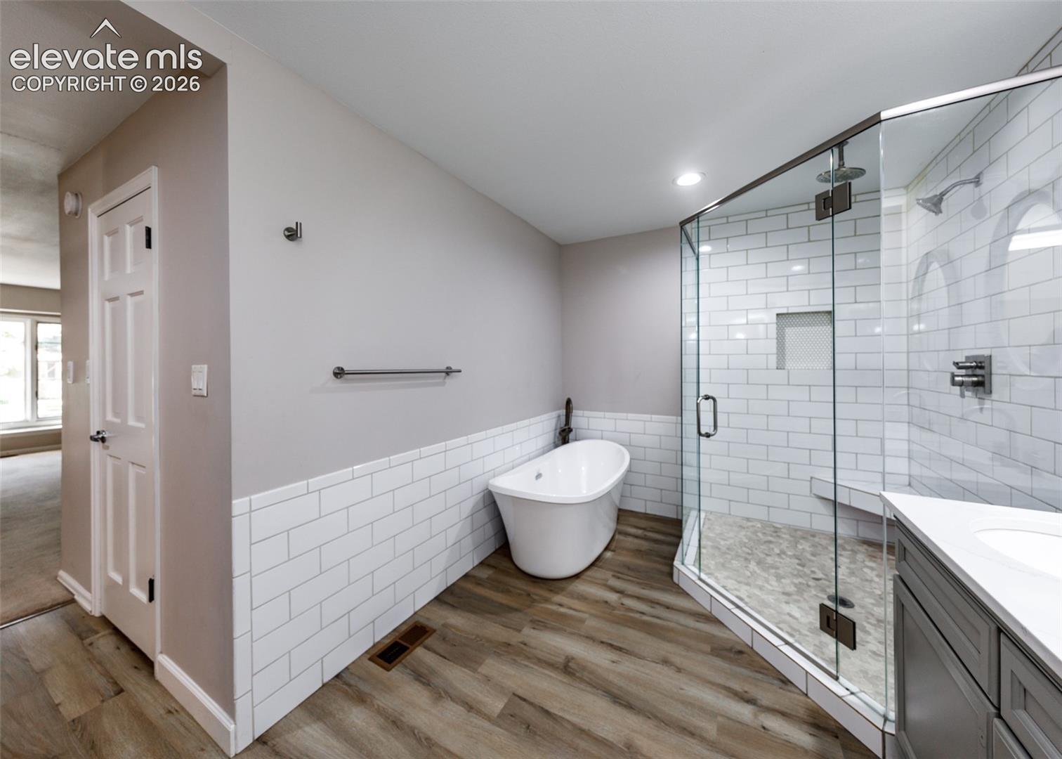 Freestanding soaking tub, walk-in shower with clear glass enclosure and tile surround, wood-finish flooring, and a single vanity with a white countertop