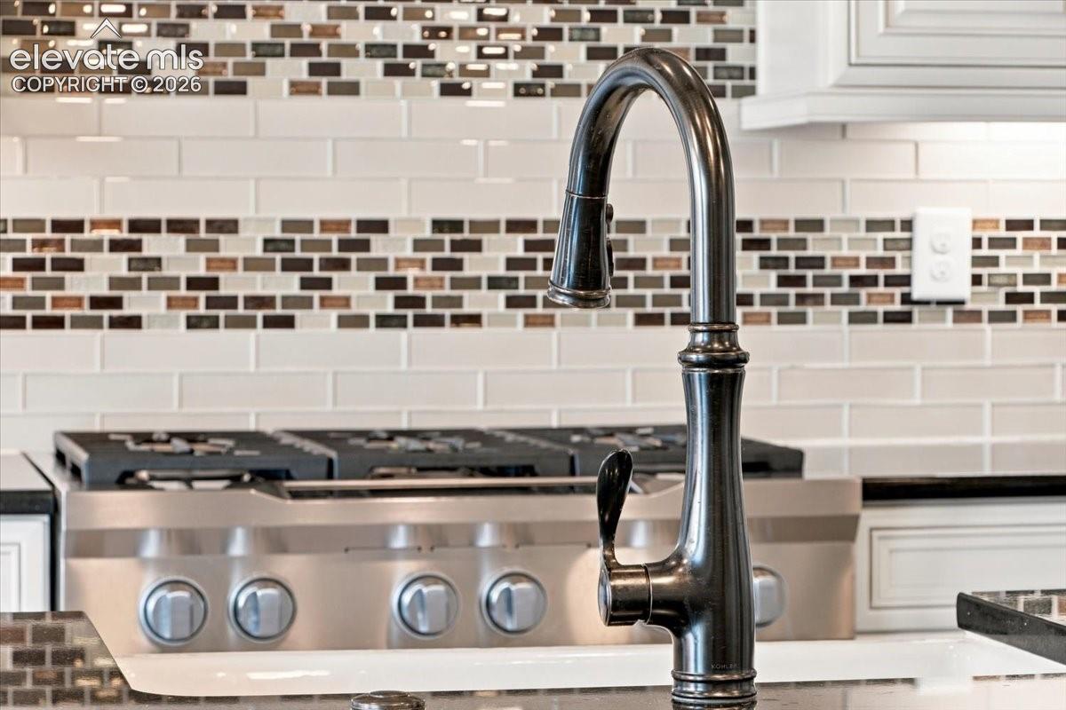 Kitchen featuring a high-arc, oil-rubbed bronze faucet, stainless steel gas range, and a multi-toned glass tile backsplash with white cabinetry