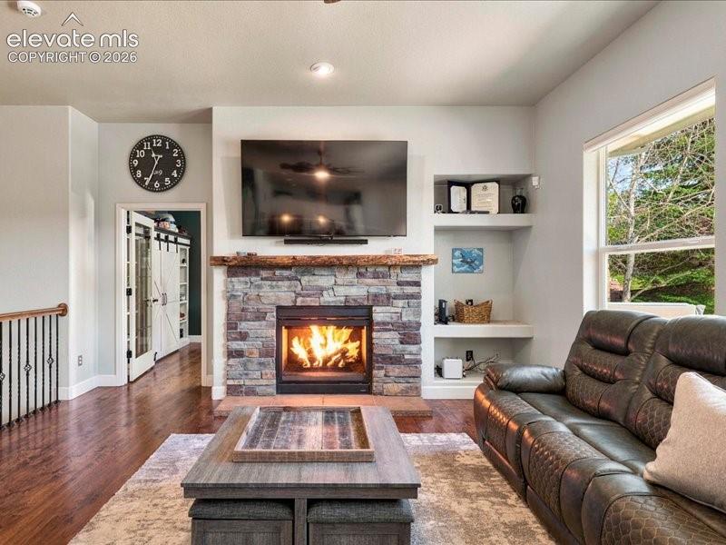 Stone-clad fireplace with wood mantel, built-in shelving, wood-finish flooring, and a large window