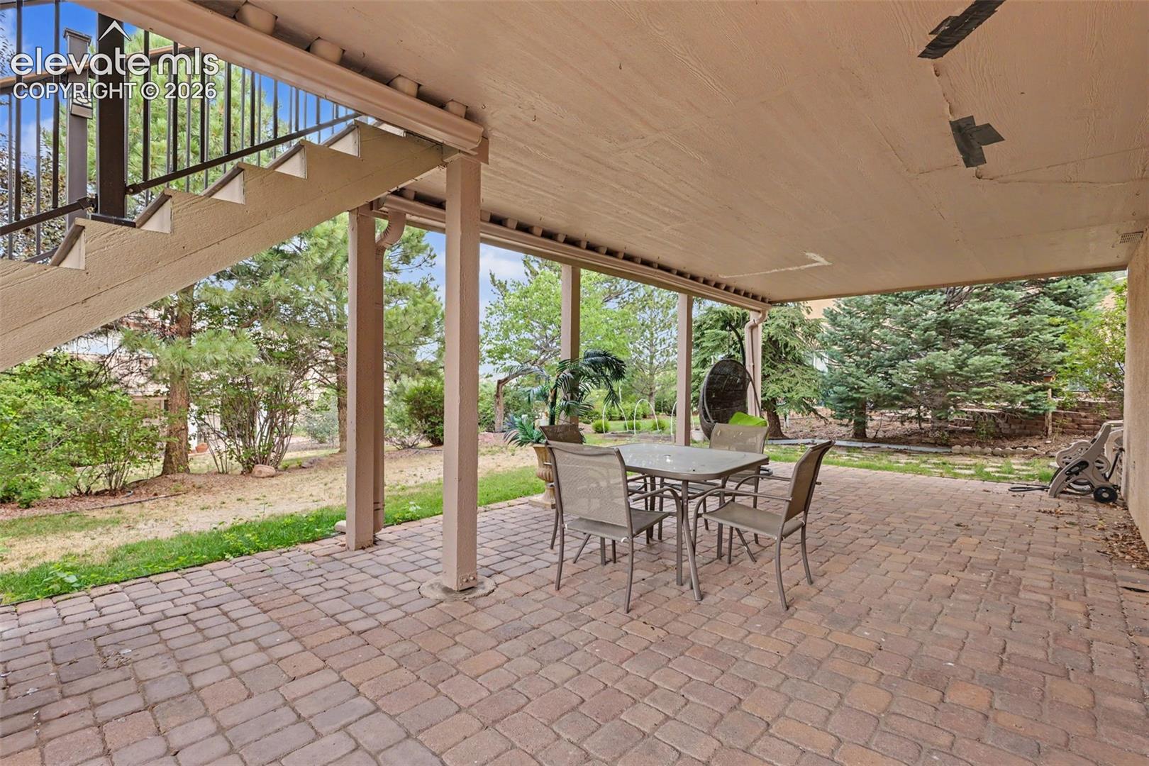 Covered outdoor patio featuring brick paver flooring, structural support columns, and an exterior staircase with black metal railings