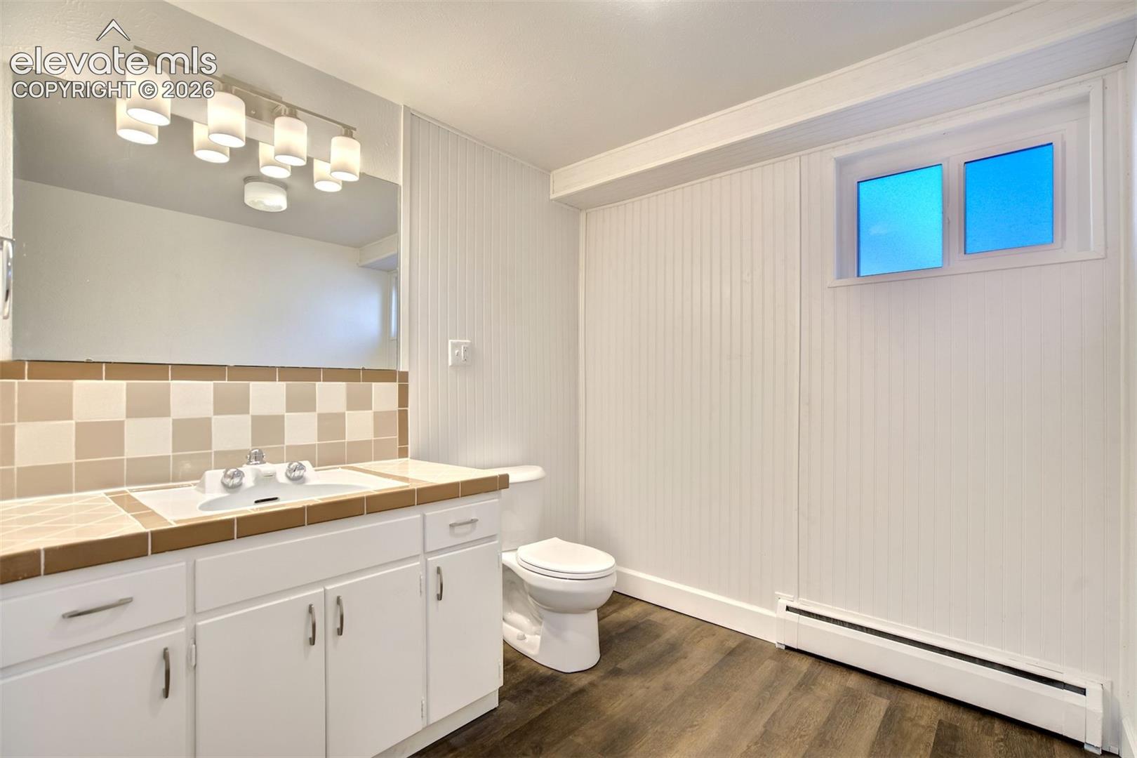 Vanity featuring white cabinetry, a ceramic tiled countertop with integrated sink, wood-finish flooring, beadboard wall paneling, and a multi-bulb light fixture