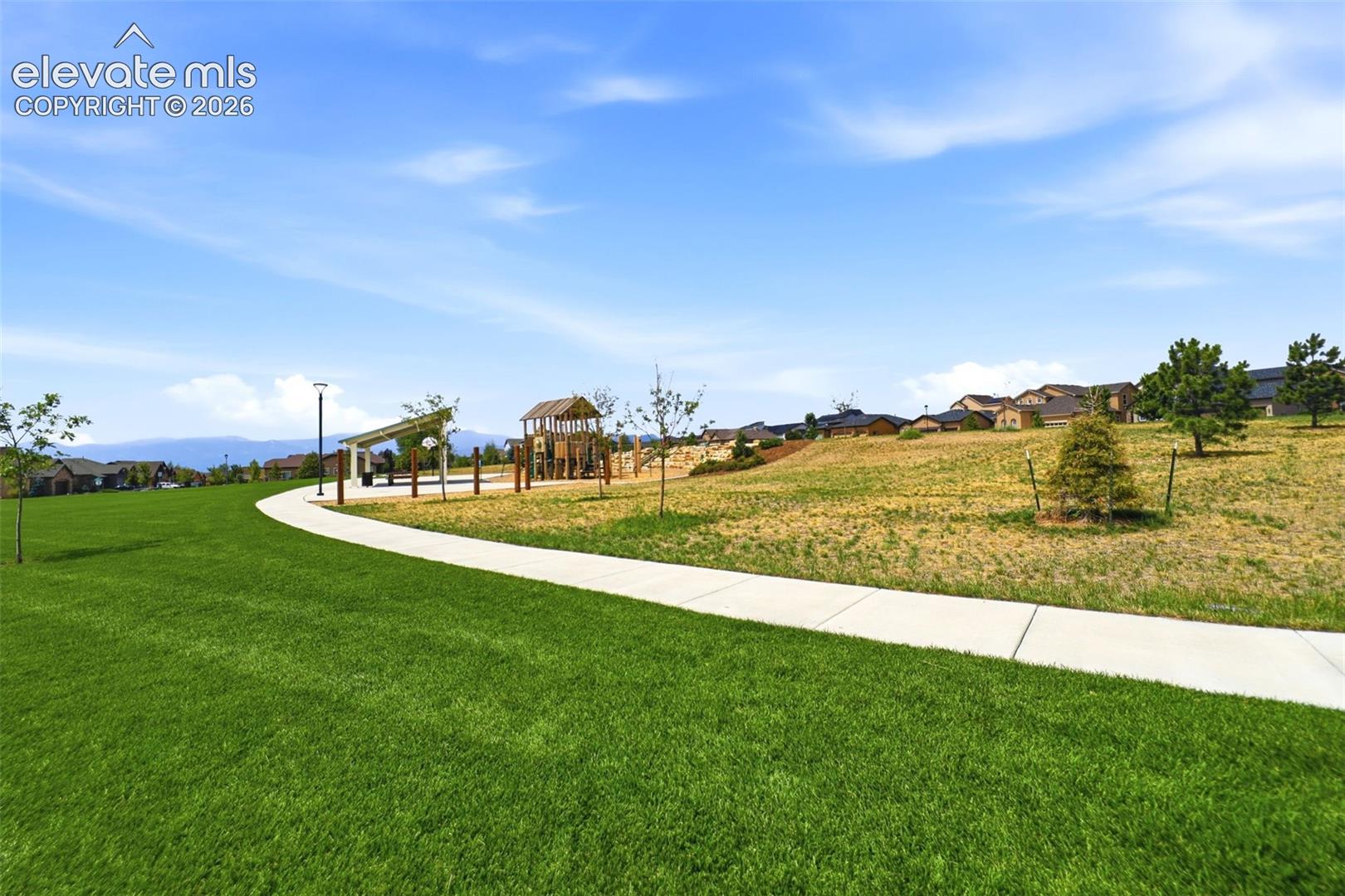 Expansive green space featuring a paved walkway, mature trees, a covered pergola, and a playground structure