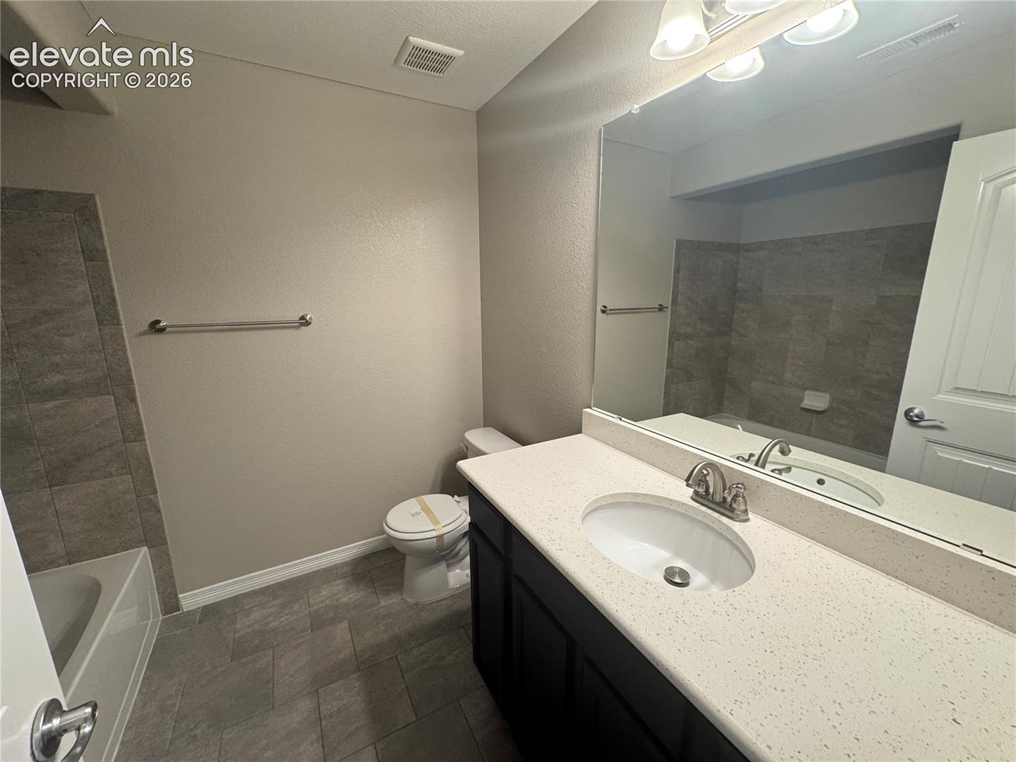 Dark-toned cabinetry topped with a light-colored solid surface vanity, integrated oval sink, full-width wall mirror, and neutral-toned ceramic tile flooring extending to the shower surround