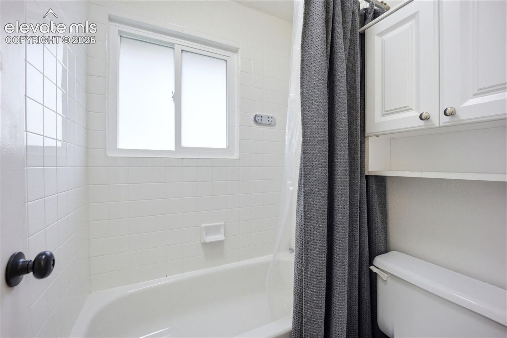 White tile tub surround with integrated soap dish, sliding window, white wall-mounted upper cabinetry, and matte black hardware