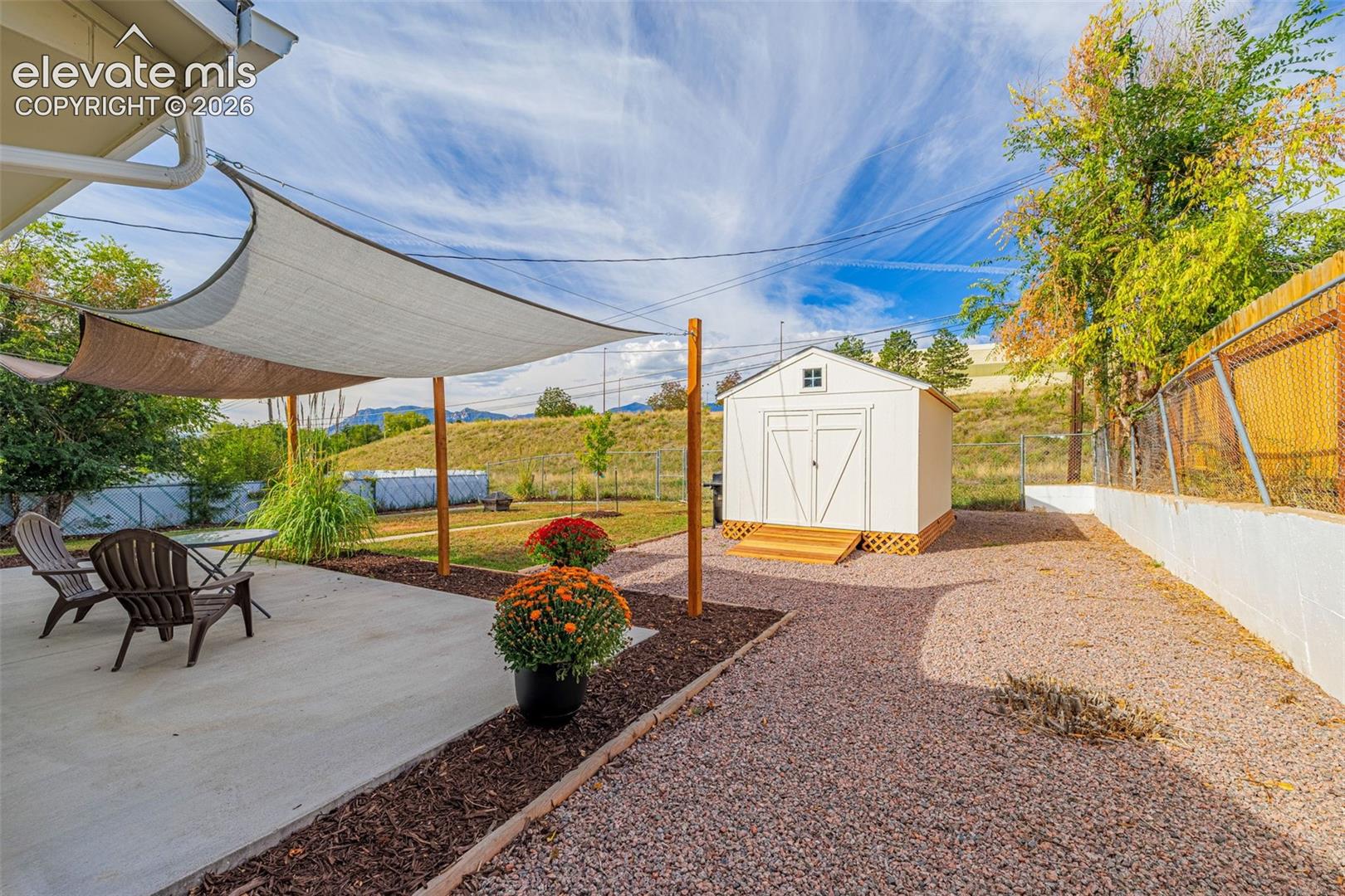 Concrete patio area, shade sail, gravel landscaping, white storage shed with wood steps, and a retaining wall