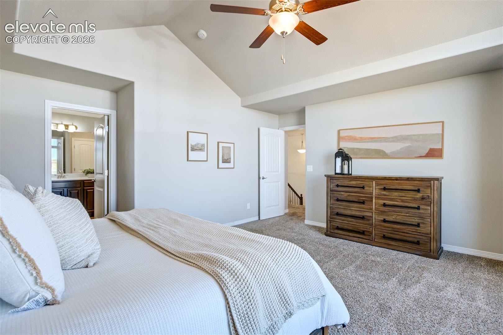 Vaulted ceiling, ceiling fan with integrated lighting, carpeted flooring, and a doorway to a bathroom with a vanity