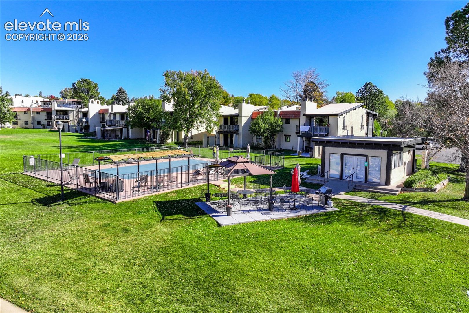Community pool enclosed by wrought iron fencing, paved patio space, covered gazebo structure, and expansive manicured lawn