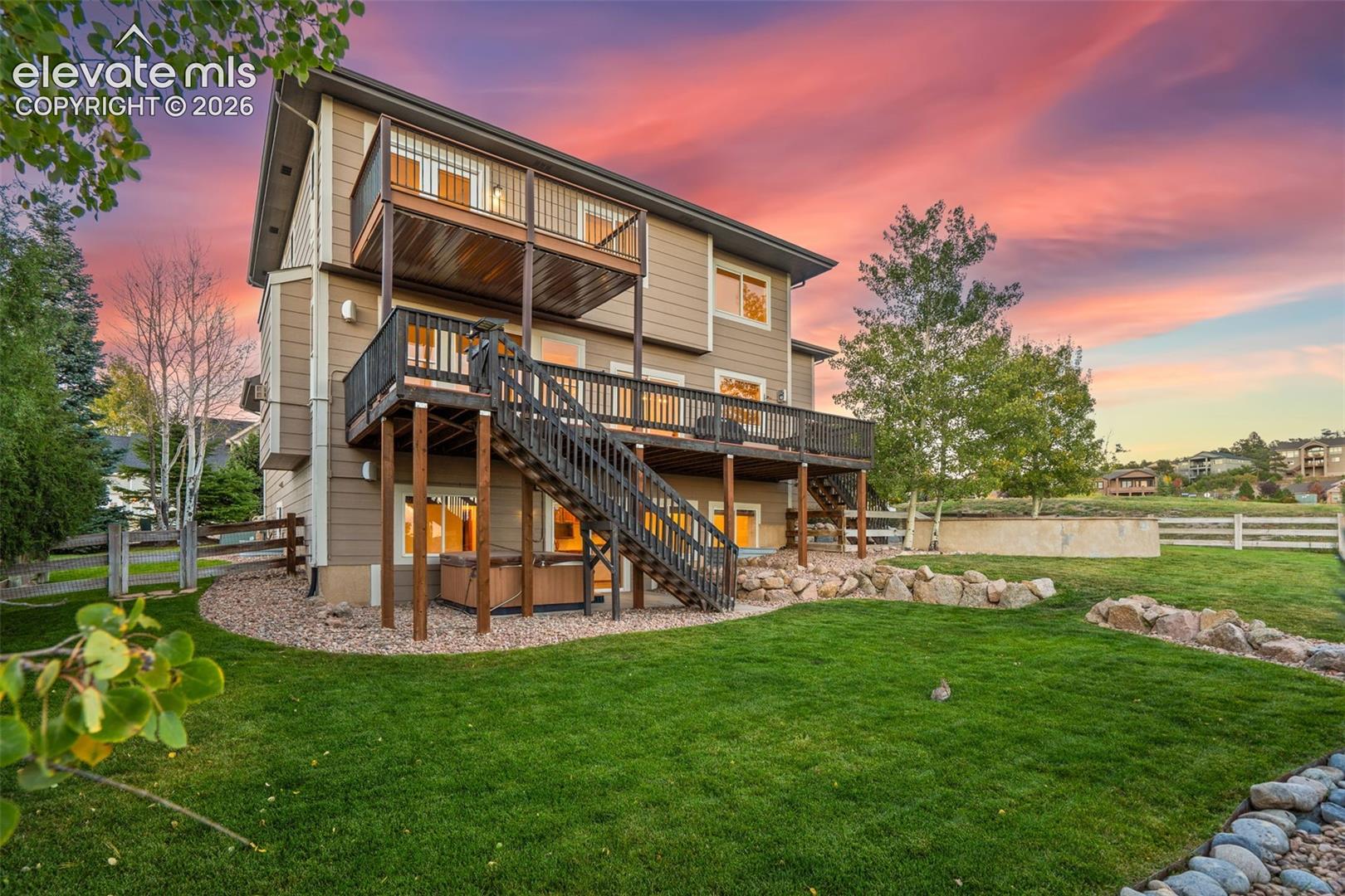 Multi-level exterior featuring wood siding, tiered elevated wooden decking, exterior staircase, and rock landscaping set against a backdrop of mature trees