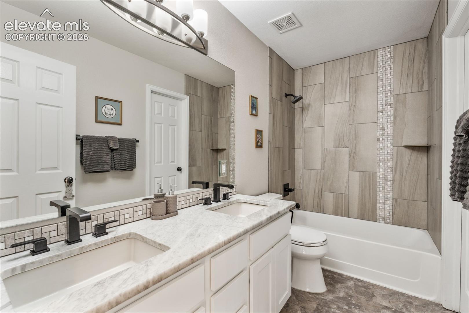 Bathroom featuring a double-sink vanity with stone countertops, matte black plumbing fixtures, oversized tile shower surround with glass mosaic accents, and an oval vanity light fixture