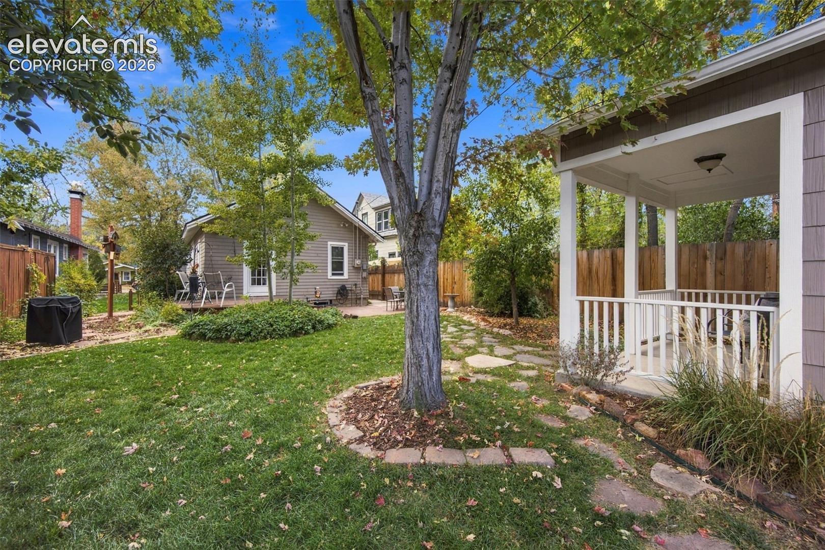 Backyard featuring a covered porch with white railing, flagstone pathway, mature trees, and wood-fence perimeter