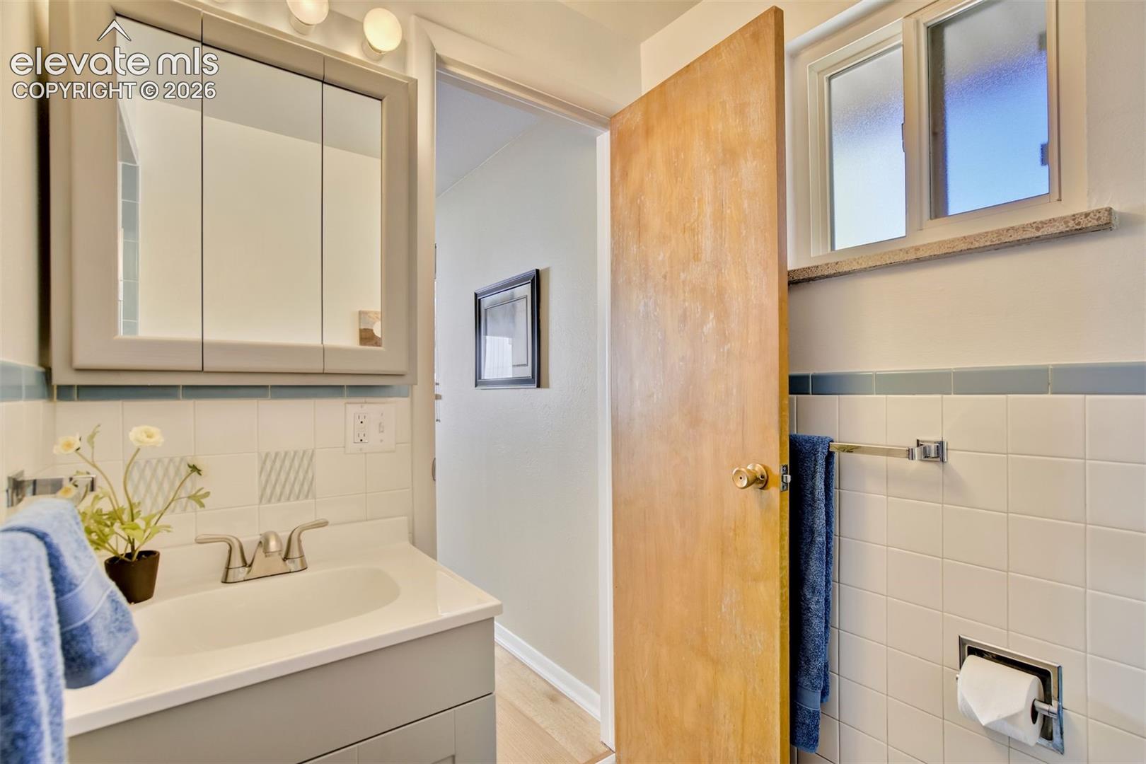 Neutral-toned vanity with an integrated basin, mirrored medicine cabinet, wall-mounted light fixture, white tile backsplash with blue accent trim, and window with stone sill