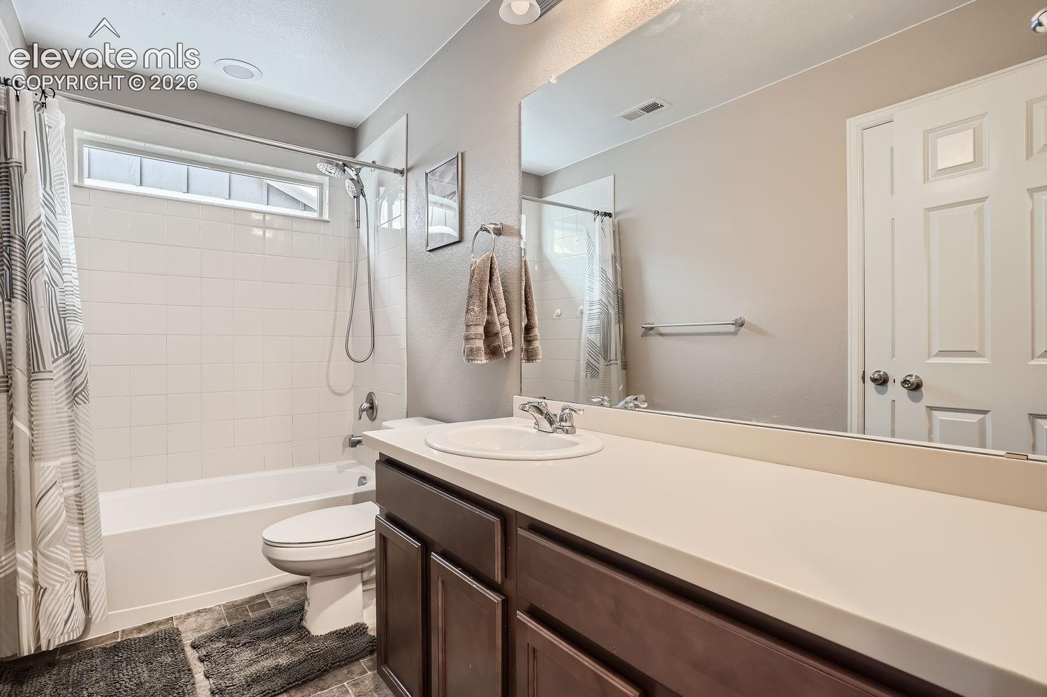 Bathroom featuring a vanity with dark-finish cabinetry, light-toned solid surface countertop, integrated sink, and full-width vanity mirror, paired with a shower-tub combination featuring a horizontal transom window and wall-mounted shower fixture
