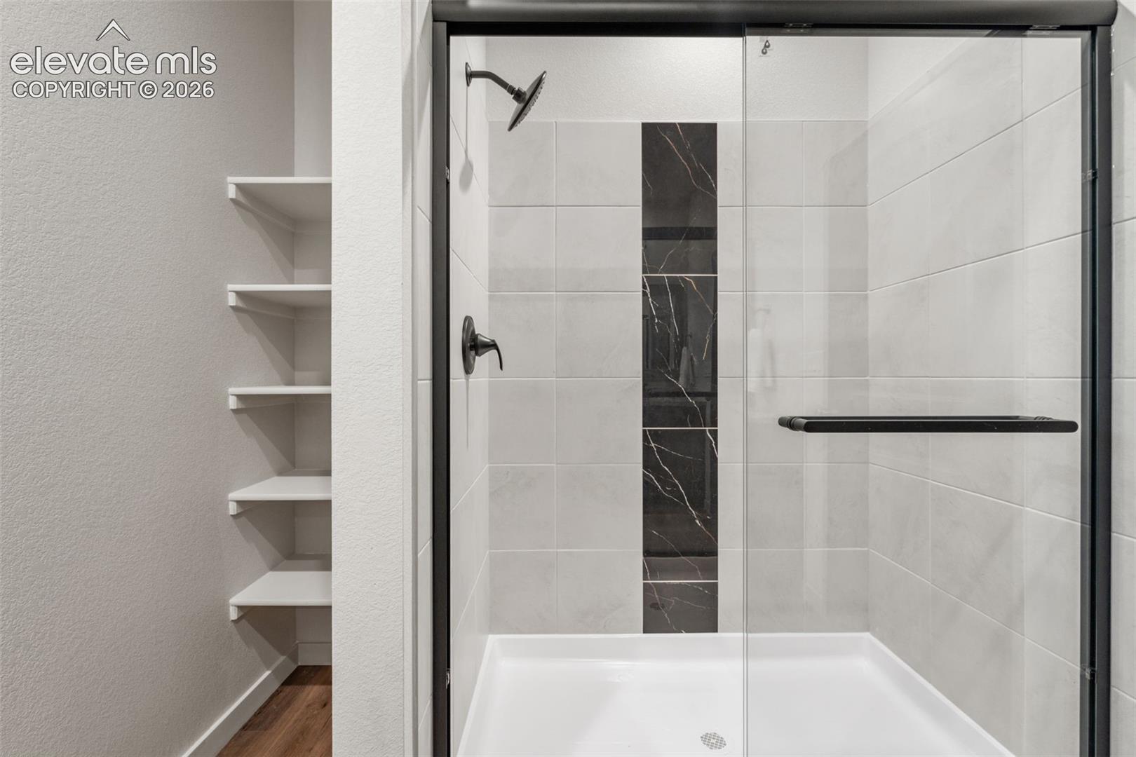 Walk-in shower featuring large-format wall tile, decorative dark stone-finish accent tile, a glass door with a matte black frame, and a matching matte black shower fixture, complemented by built-in wall shelving