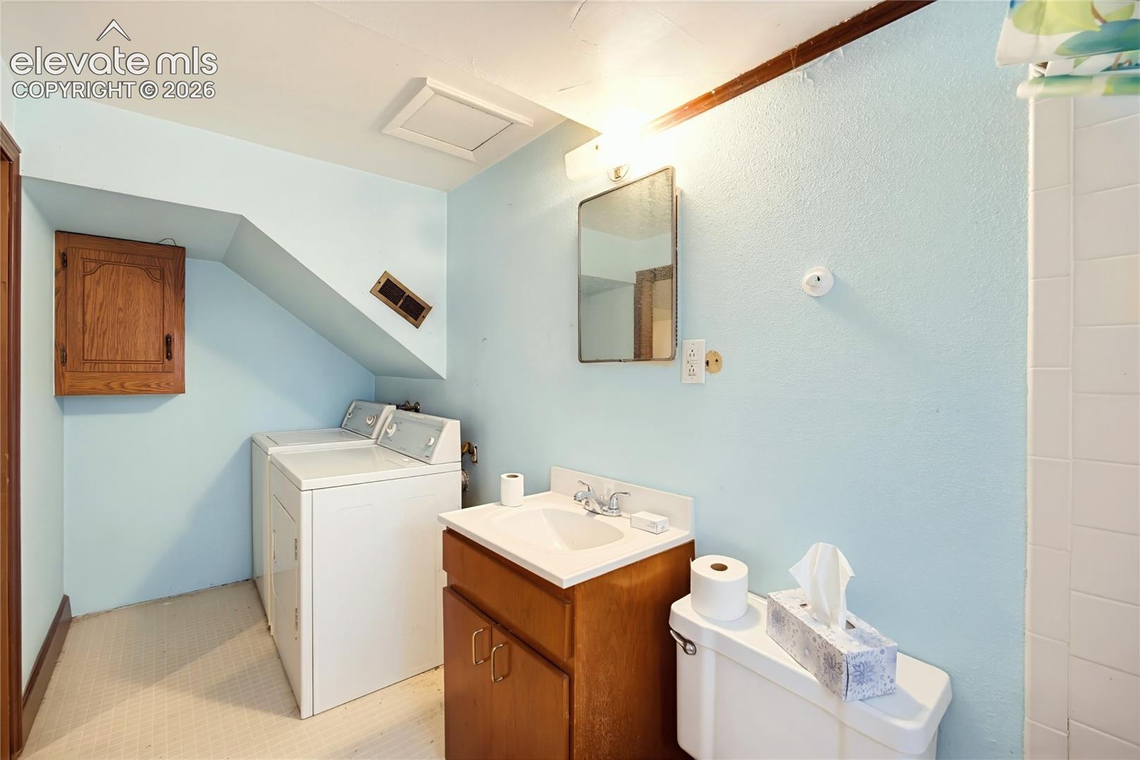 Built-in laundry appliances, wood-finish vanity with an integrated sink, wall-mounted mirror, and tiled shower surround