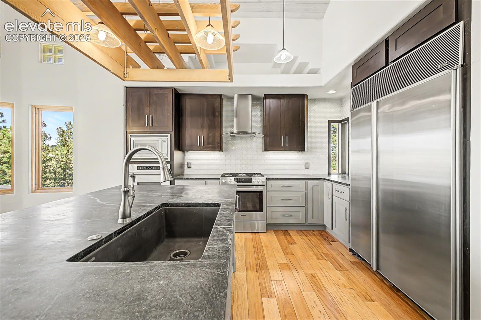 Kitchen featuring a large island with an undermount sink and gooseneck faucet, stainless steel appliances, wood-finish flooring, white subway tile backsplash, and vaulted ceilings with exposed beams