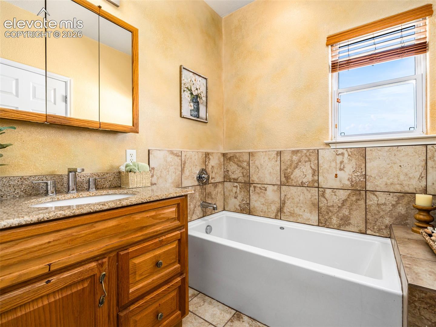Built-in bathtub with tiled surround, wood-finish vanity with granite countertop, and integrated sink