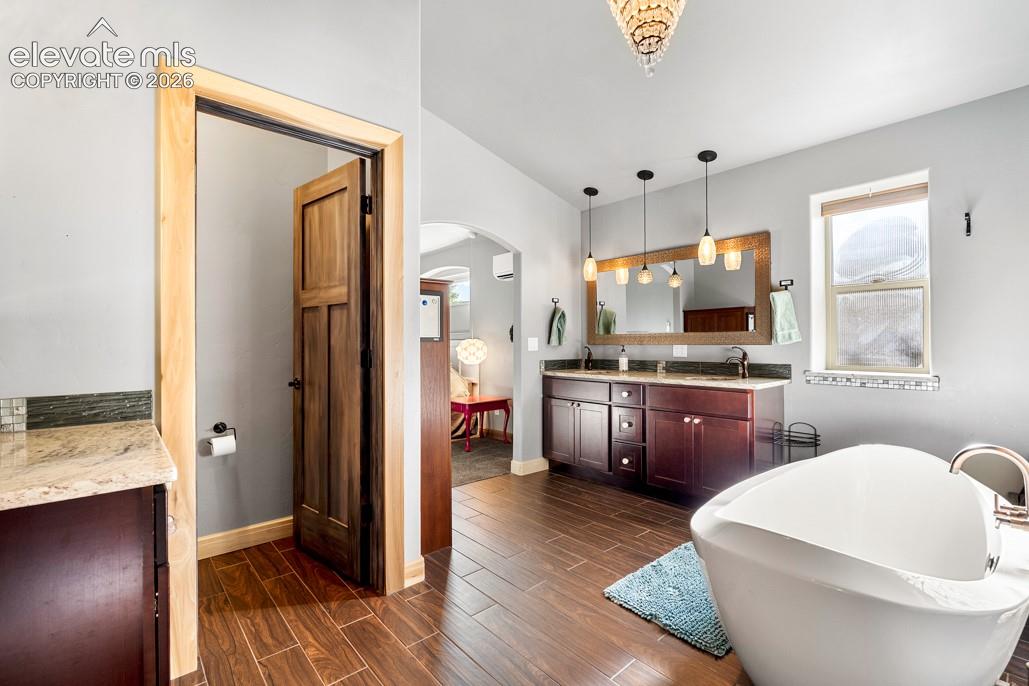 Freestanding soaking tub, wood-finish flooring, dual vanity with undermount sinks, granite-look countertop, and pendant lighting