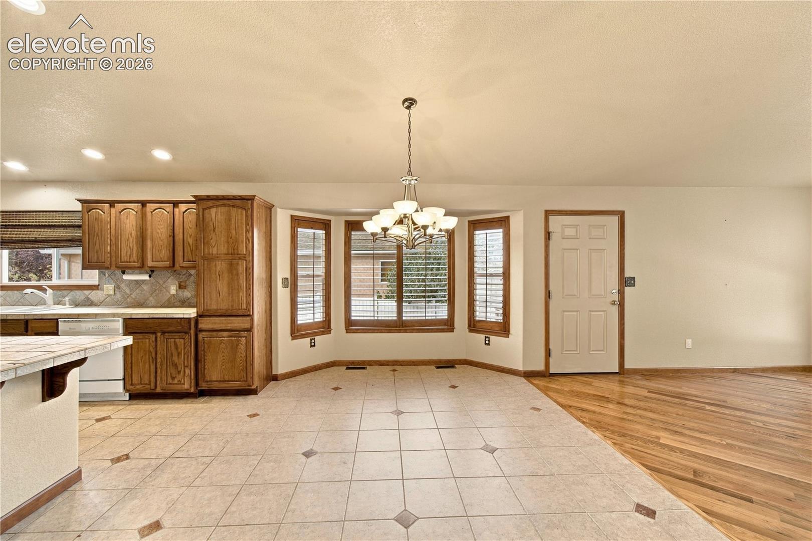 Kitchen featuring wood cabinetry, ceramic tile flooring with decorative inserts, a bay window with plantation shutters, a multi-light chandelier, and wood-finish flooring