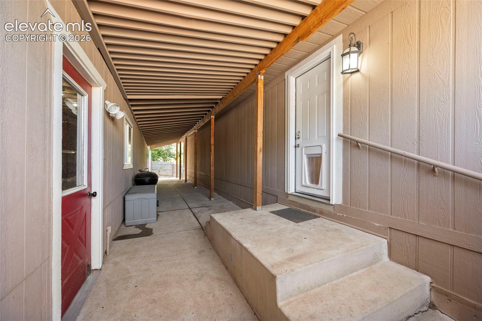 Covered exterior walkway featuring vertical wood-composite siding, concrete flooring, attached wall-mounted lantern lighting, and a raised concrete entry landing with a handrail