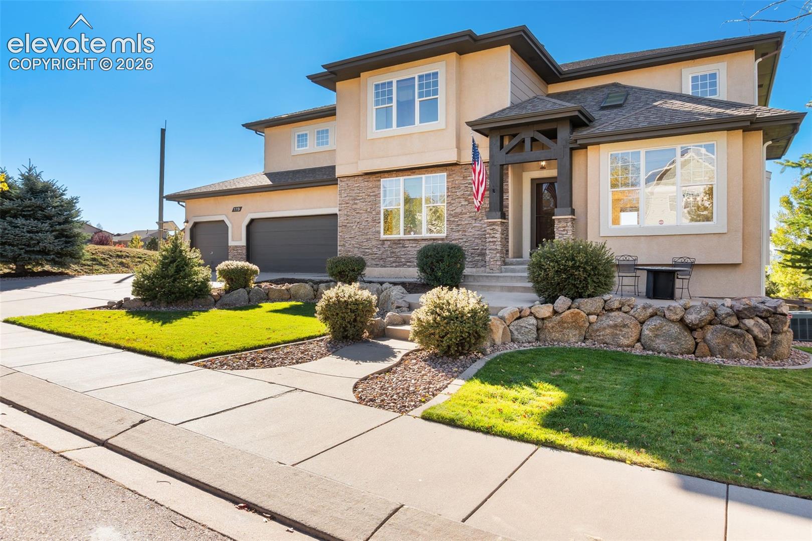 Two-story exterior featuring a stone veneer facade, stucco siding, a covered entryway with decorative timber truss detailing, and an attached three-car garage